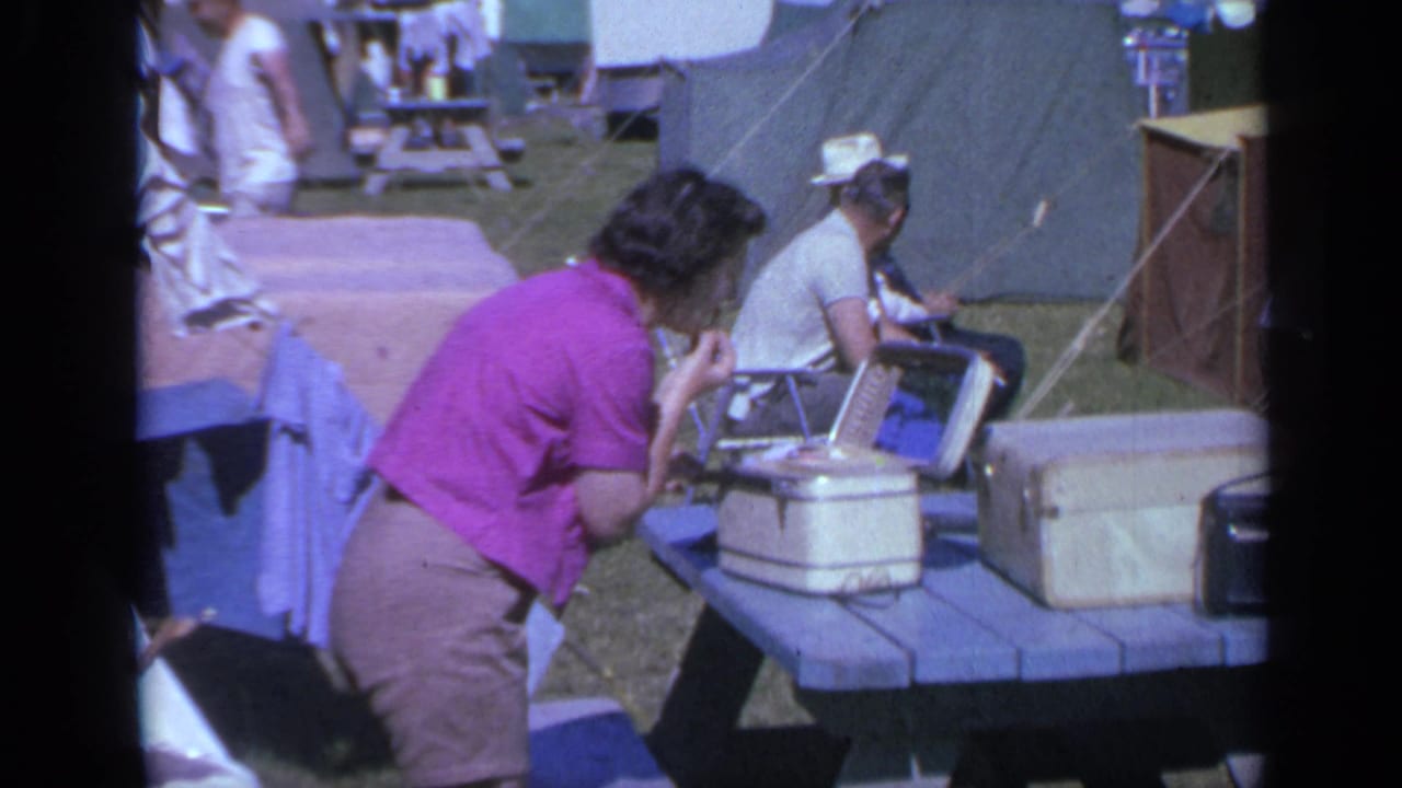 1959 Colorado campsite view with a woman in a purple shirt leaning near luggage on a picnic table as a man in a cowboy hat sits nearby.