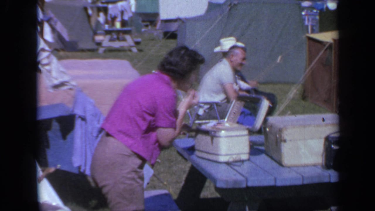 Woman bends toward an open case on a picnic table as a seated man smiles nearby.