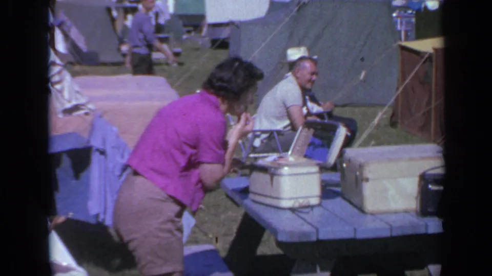Home-movie frame from a 1959 Colorado campsite showing a woman leaning over a table with luggage while a man sits near tents.