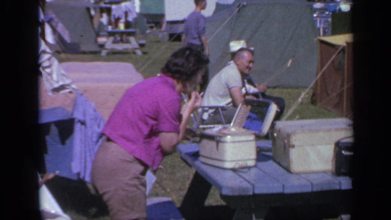 1959 Colorado campground frame with a woman beside table luggage, a seated man in a cowboy hat, tents, and laundry in the background.