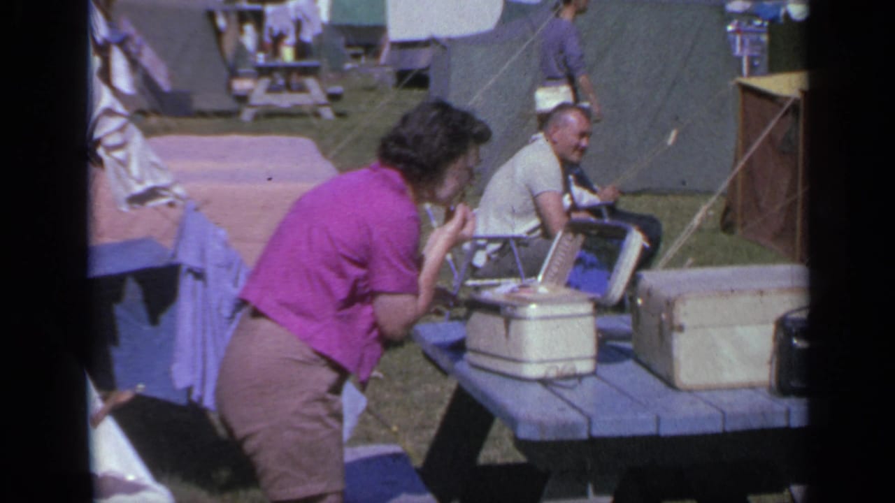 Woman leans over a picnic table using a mirror while a seated man smiles near tents and coolers.