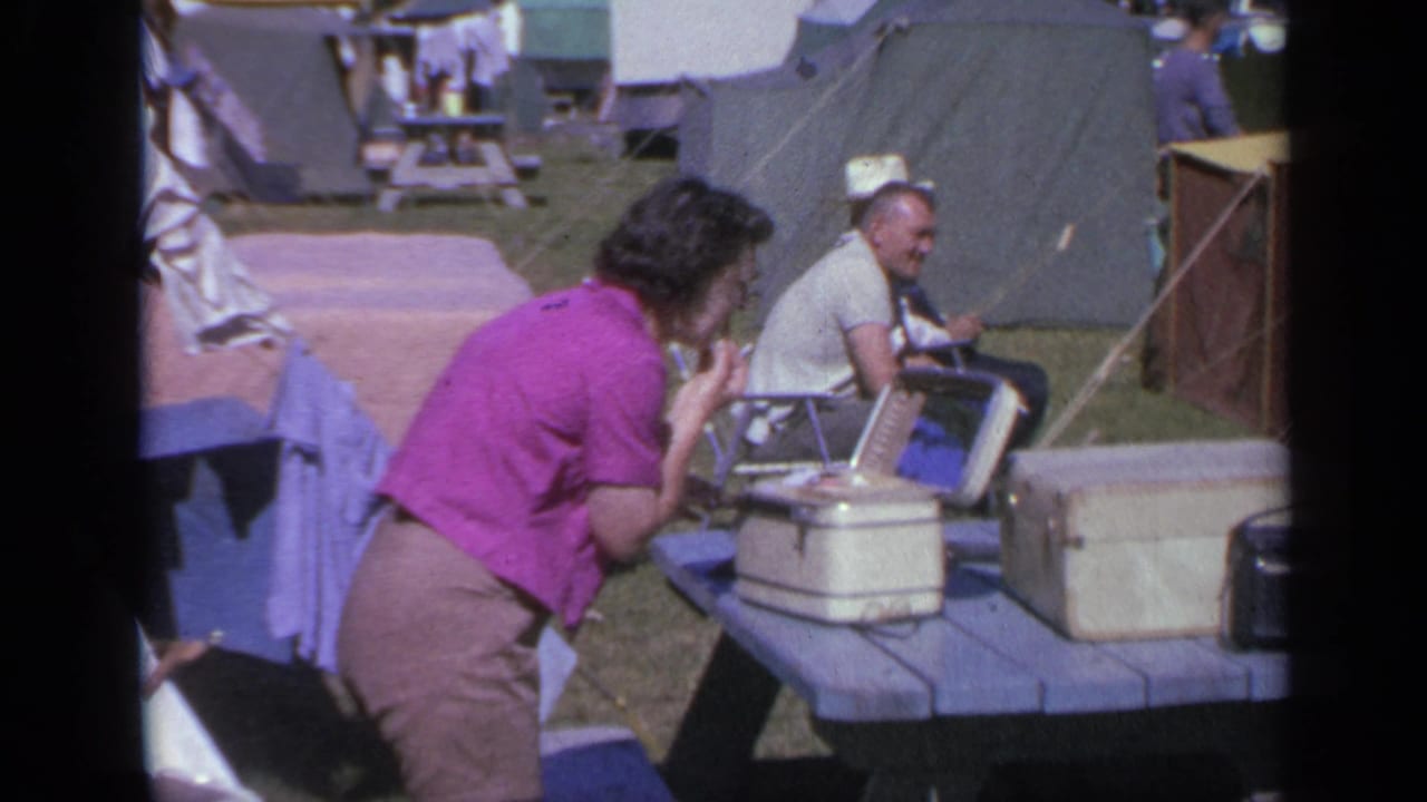 A woman uses a mirror at a campsite picnic table in Colorado, with a seated man, coolers, tents, and laundry visible around her.