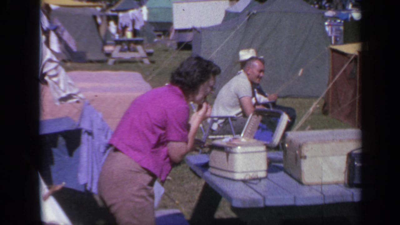 At a 1959 Colorado campsite, a woman appears to apply makeup near a mirror on a picnic table, with tents and coolers behind her.