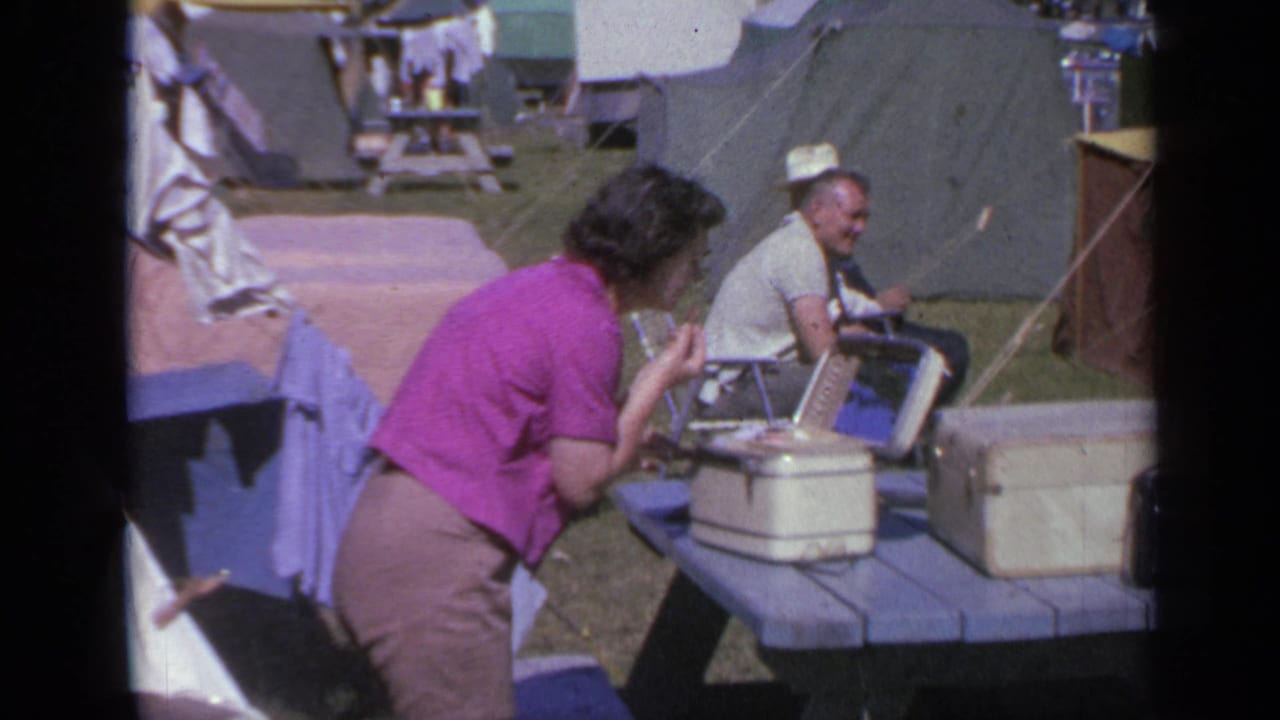 Woman in pink bends over a campsite picnic table while a seated man in a hat watches nearby.
