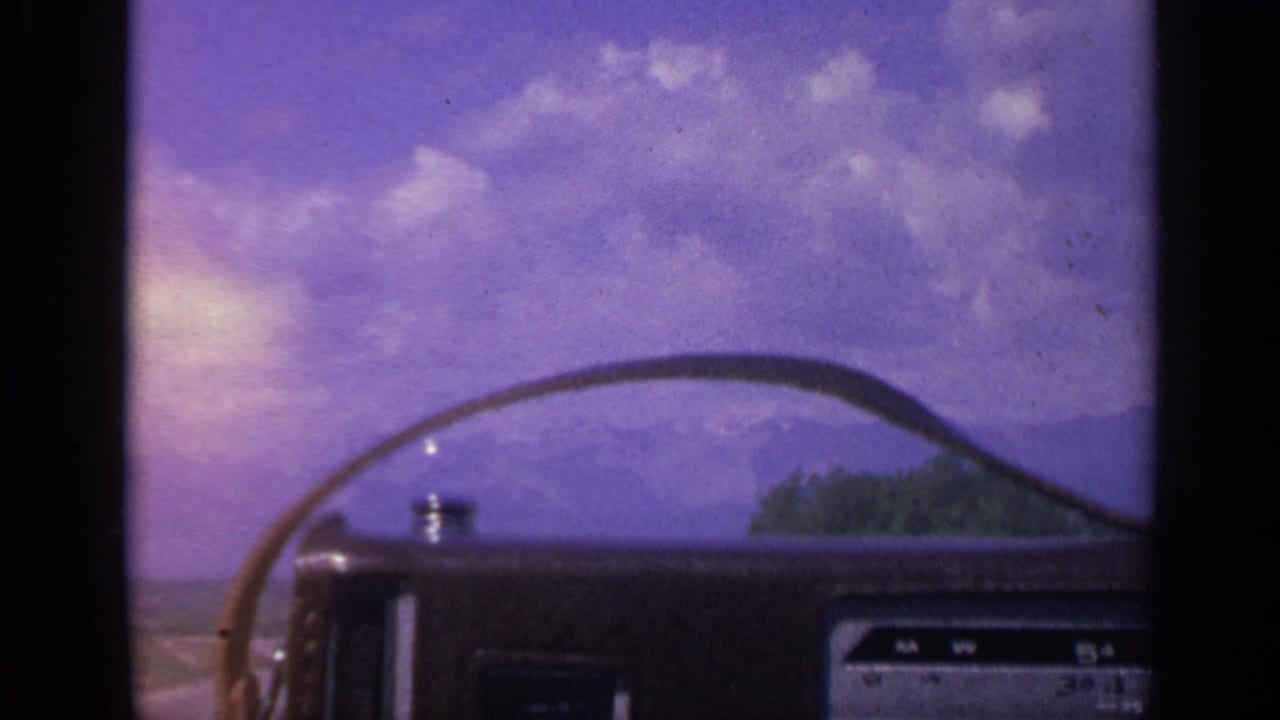 1959 Colorado travel shot from inside a vehicle, with windshield frame, dashboard detail, road, hills, and roadside trees.
