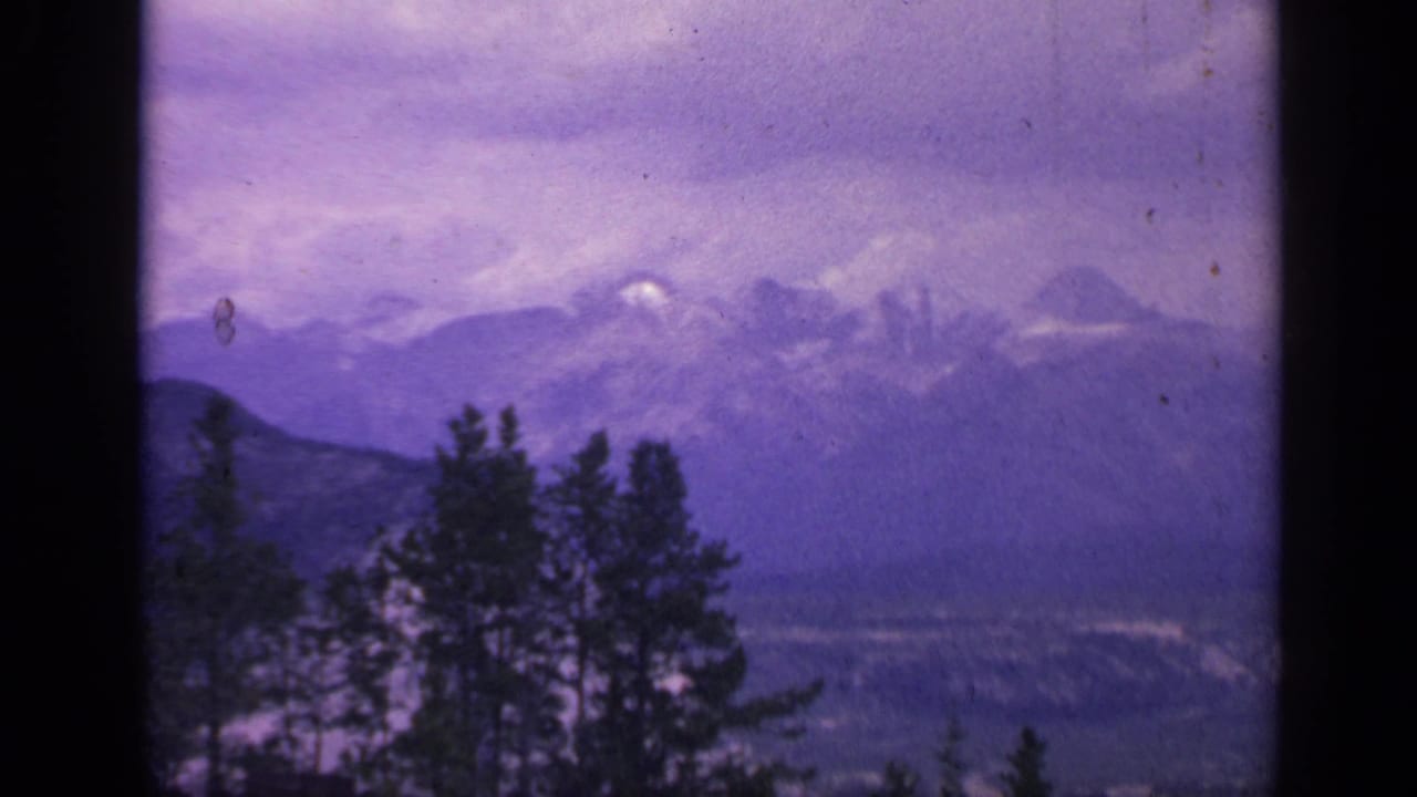 1959 Colorado mountain view with foreground evergreens, hazy peaks, and a violet color-shifted storm sky.