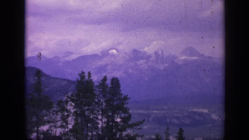 1959 Colorado mountain view with foreground evergreens, hazy peaks, and a violet color-shifted storm sky.