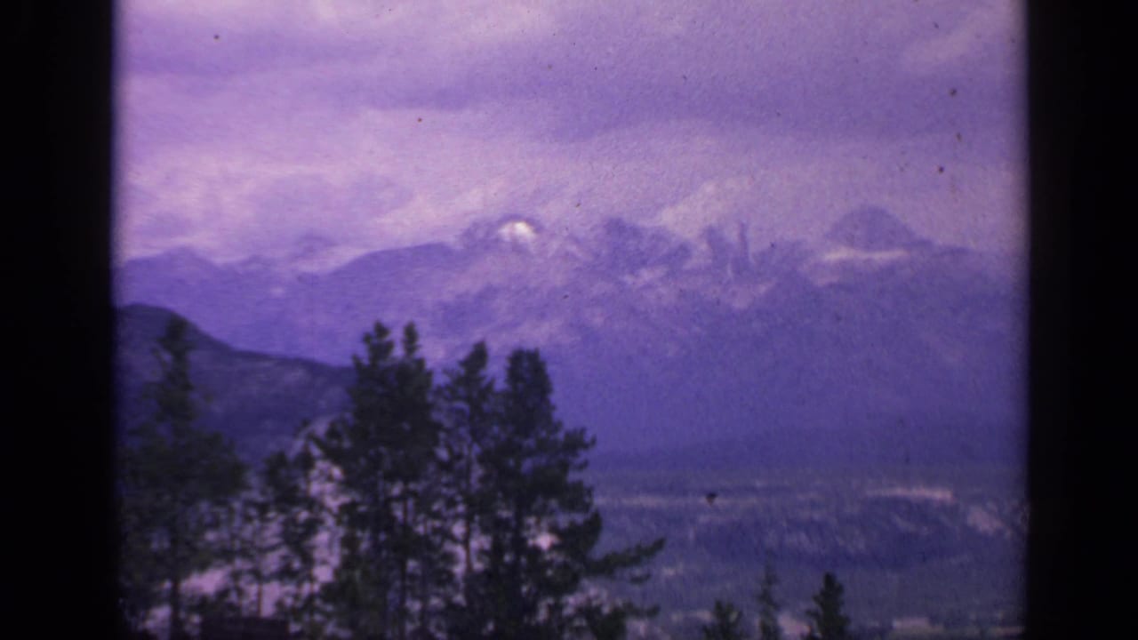 1959 Colorado 8mm landscape frame with dark evergreens below hazy snow-capped peaks and a purple cloudy sky.