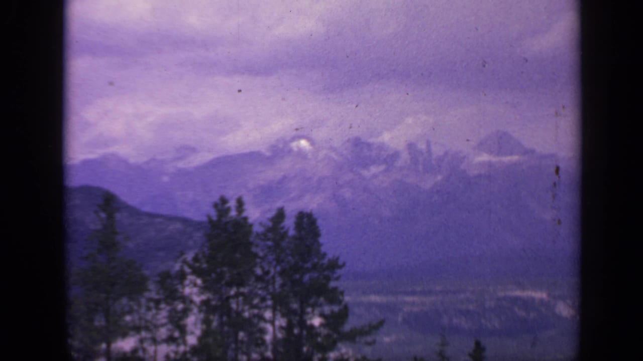 1959 Colorado scenic frame showing evergreen silhouettes, distant mountain slopes, snow patches, and a purple-gray sky.