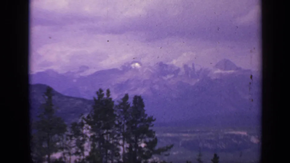 1959 Colorado scenic frame showing evergreen silhouettes, distant mountain slopes, snow patches, and a purple-gray sky.