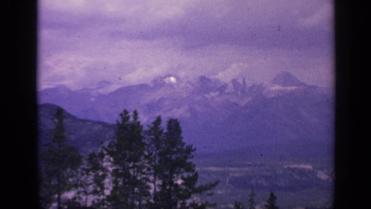 1959 Colorado home-movie frame of snow-dusted mountains, dark tree silhouettes, and a hazy valley below.