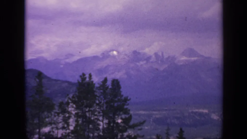 1959 Colorado home-movie frame of snow-dusted mountains, dark tree silhouettes, and a hazy valley below.