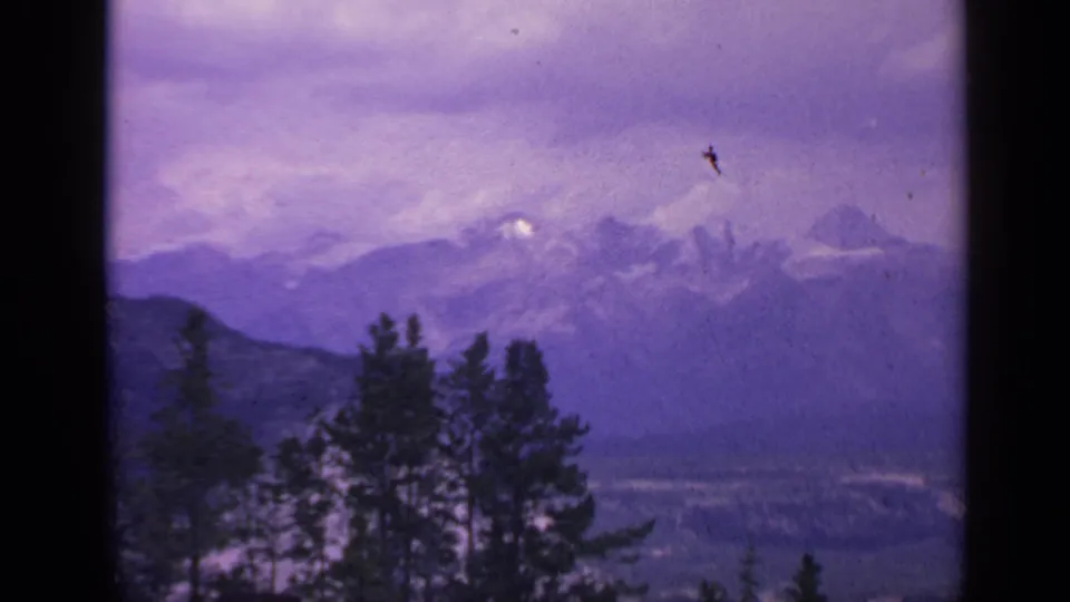 1959 Colorado 8mm frame with evergreen treetops, hazy snow-capped mountains, and a small dark speck crossing the purple sky.