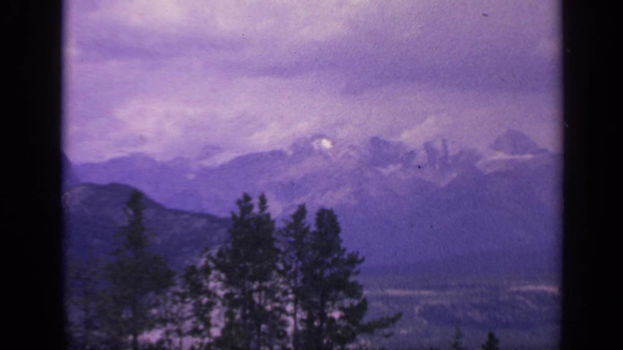 1959 Colorado 8mm frame with soft evergreen silhouettes, distant snow-marked mountains, and purple color shift across the cloudy sky.