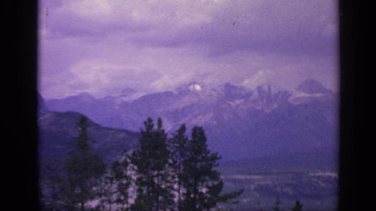 1959 Colorado 8mm frame showing distant snow-dusted ridges, a hazy sky, and dark evergreen trees in the foreground.