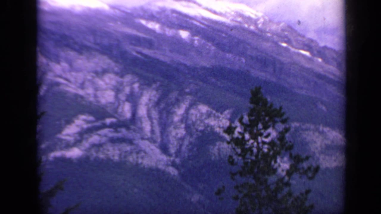 1959 Colorado 8mm frame showing a rugged snow-marked mountain face with a foreground evergreen and visible film grain.