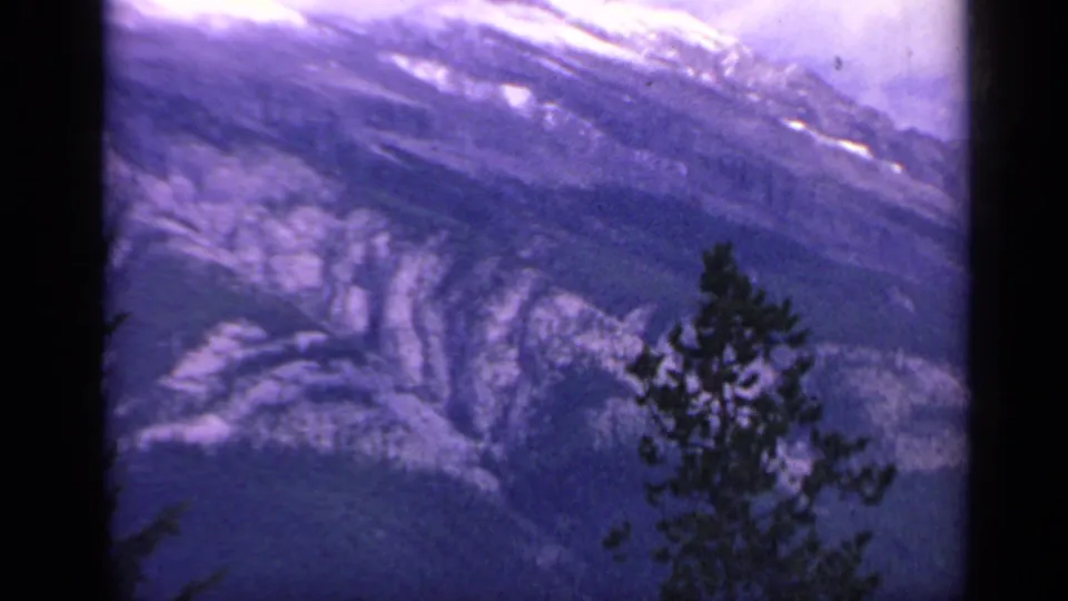 1959 Colorado 8mm frame showing a rugged snow-marked mountain face with a foreground evergreen and visible film grain.