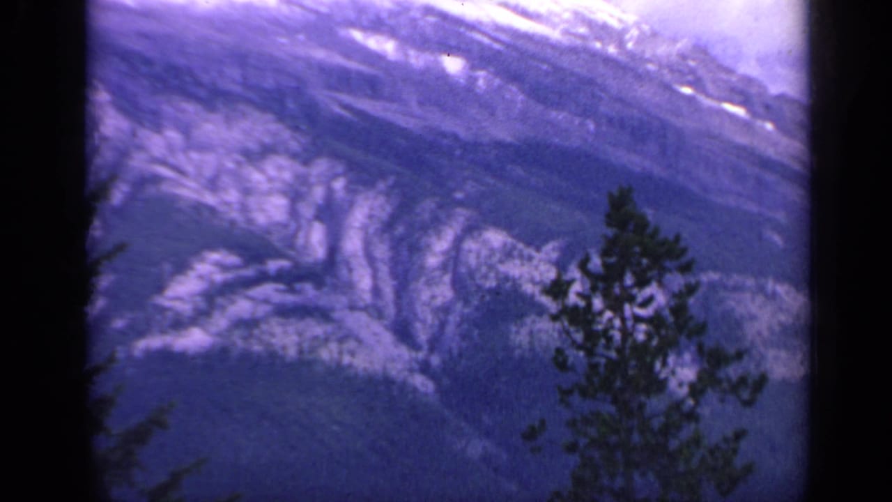 1959 Colorado 8mm frame of a snow-streaked mountain slope, foreground pines, and rounded dark frame edges.