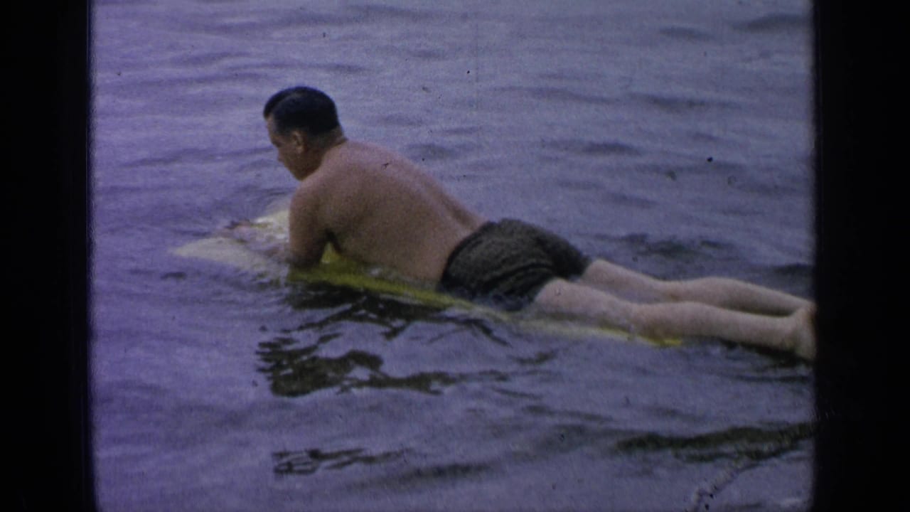 Man in swim trunks lies on a yellow board in lake water.