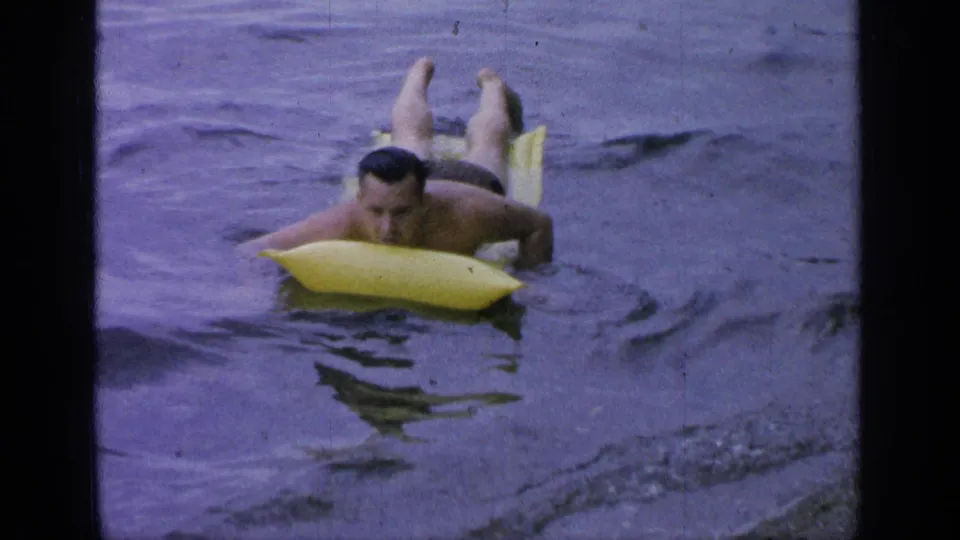 The man turns toward the camera while lying prone on the yellow board in the Greenwood Lake water.