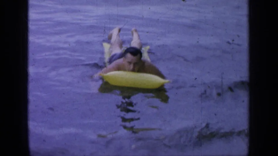 A man lies prone on a yellow inflatable raft in Greenwood Lake, with 8mm grain and dark water ripples visible.
