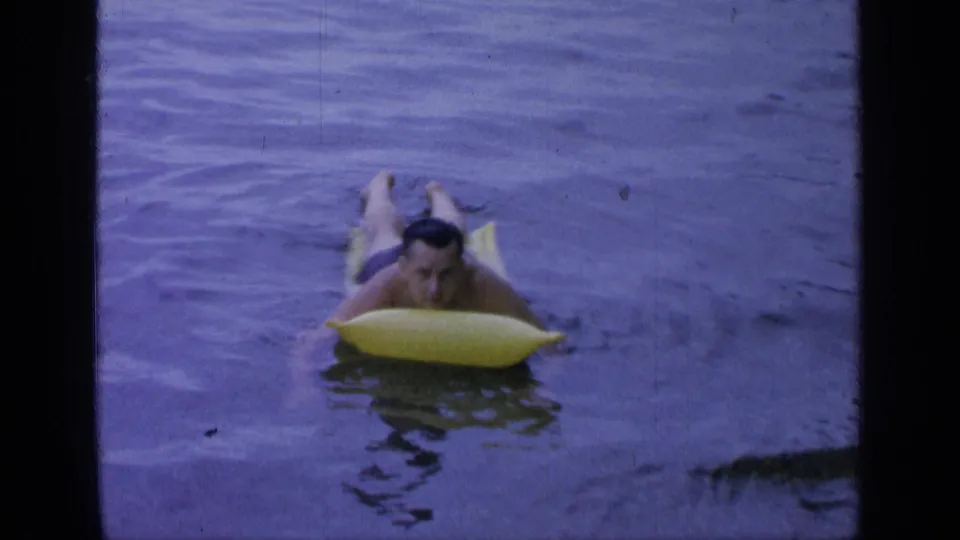 Adult man floats on a yellow inflatable raft in calm Greenwood Lake water, seen in vintage 8mm footage from 1964.