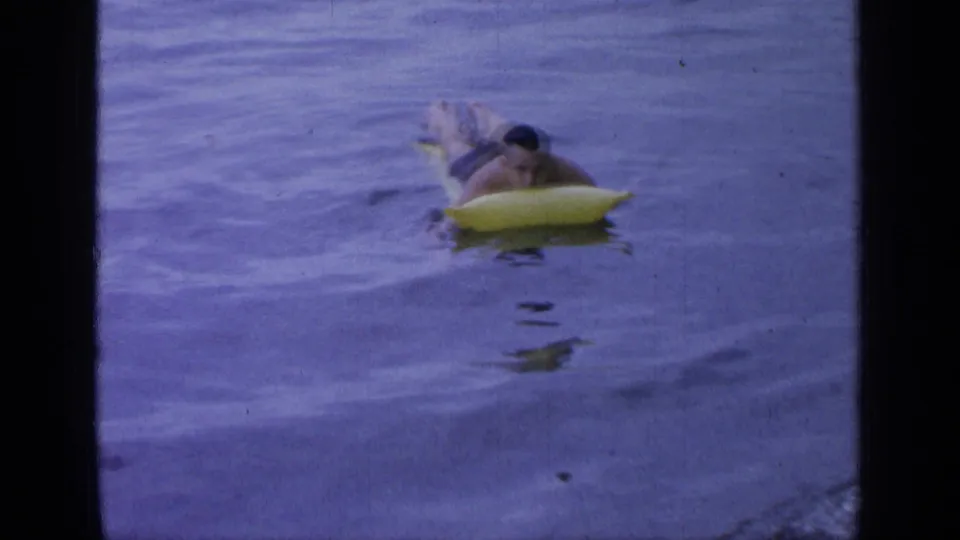 At Greenwood Lake in 1964, a man floats belly-down on a yellow inflatable raft in calm blue-purple water.