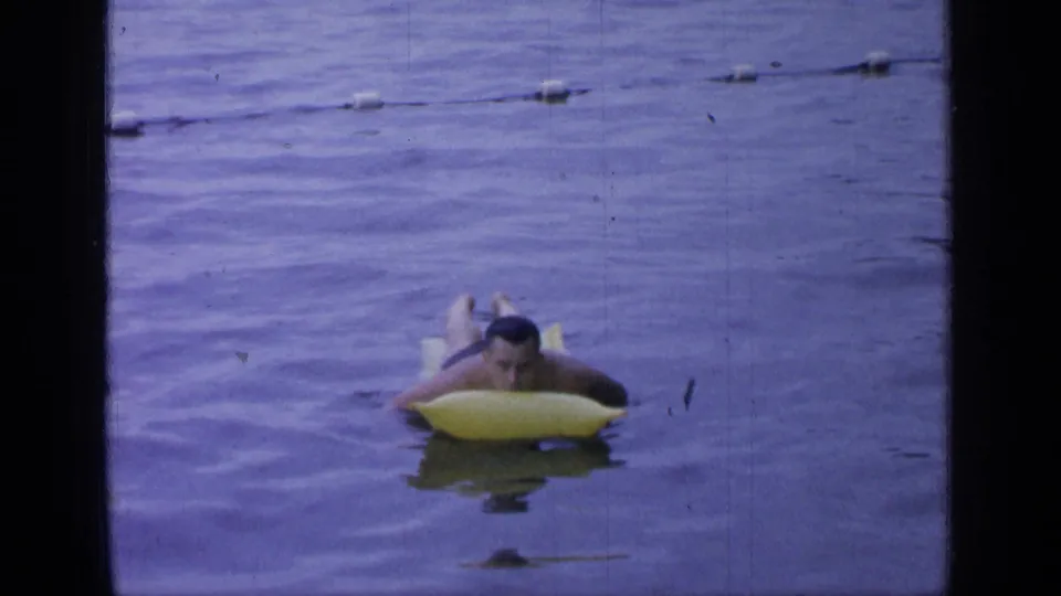 A 1964 Greenwood Lake home-movie frame shows the man approaching on the raft, partly submerged in blue-tinted water.
