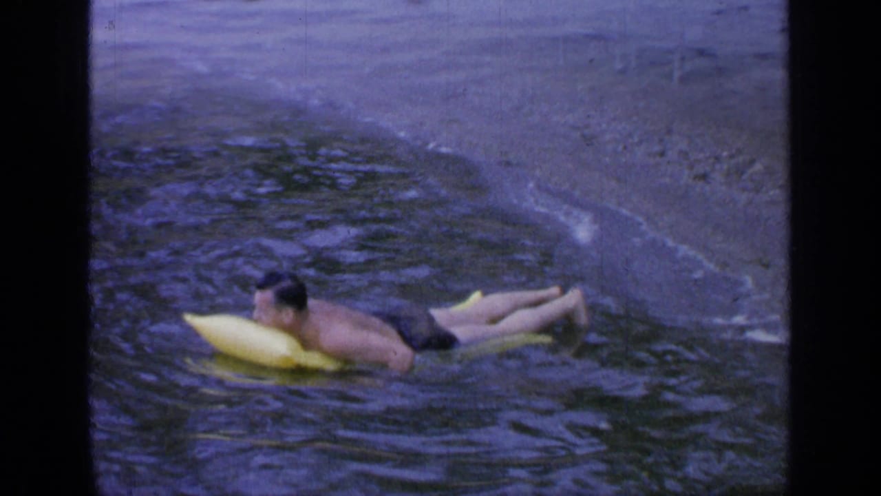 Man in black swim trunks floats on a yellow inflatable near a sandy lakeshore.