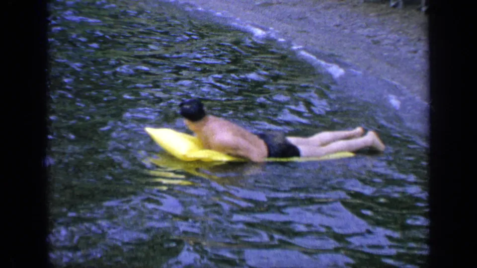 The man drifts farther across the rippled Greenwood Lake surface on a yellow inflatable cushion, with sandy shore behind him.