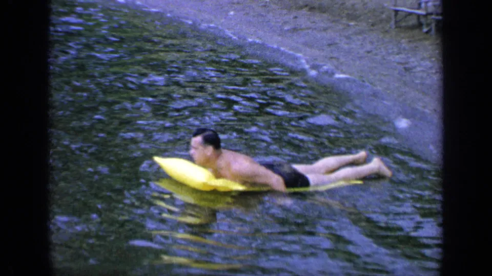 In this 1964 Greenwood Lake frame, the man turns slightly toward shore while lying on the yellow inflatable in calm water.
