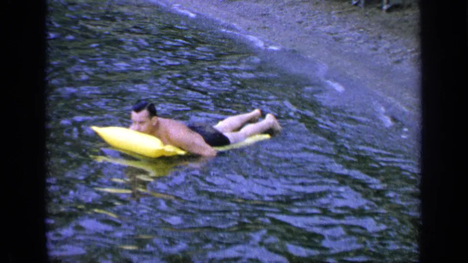 The man continues floating on the yellow cushion at Greenwood Lake, his face turned toward the camera and shore in the background.