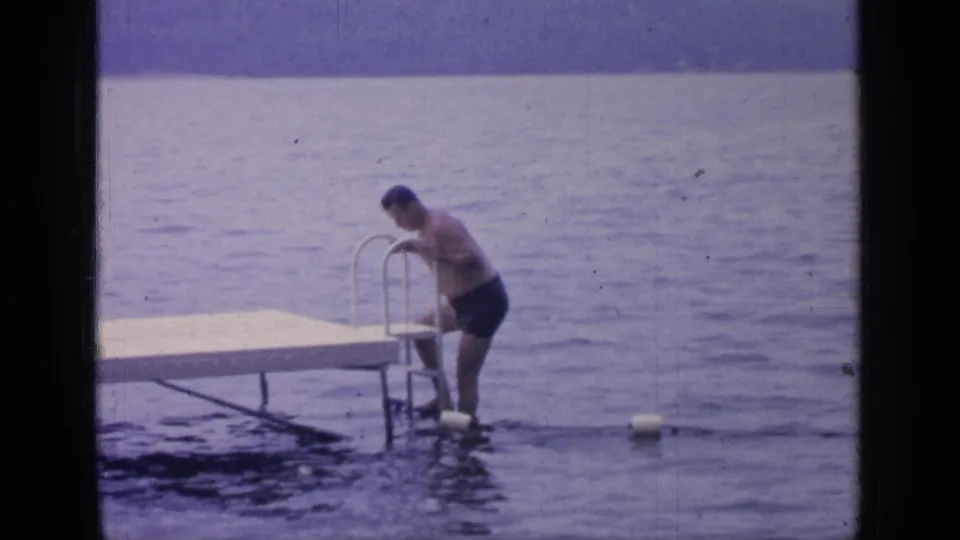1964 Greenwood Lake frame showing an adult man in blue shorts using a metal dock ladder to enter calm lake water.