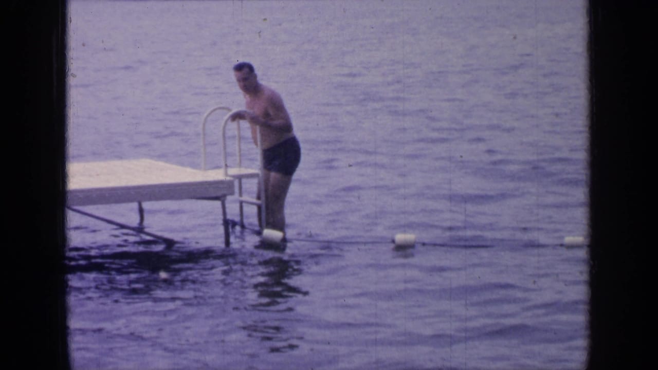 Man in dark swim trunks stands on a dock ladder with one foot near the lake water.