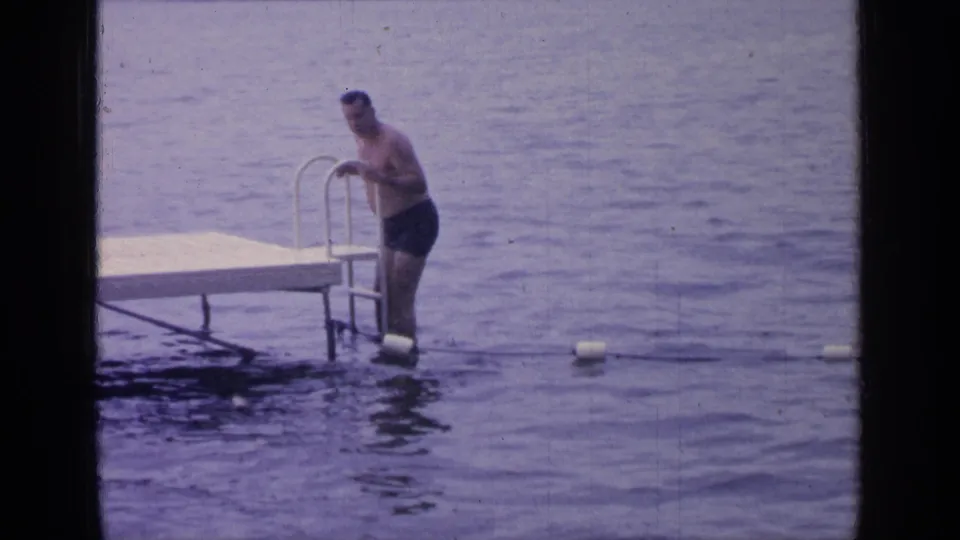 At Greenwood Lake in 1964, a man in dark swim trunks pauses on a metal ladder beside a wooden dock as he tests the water.