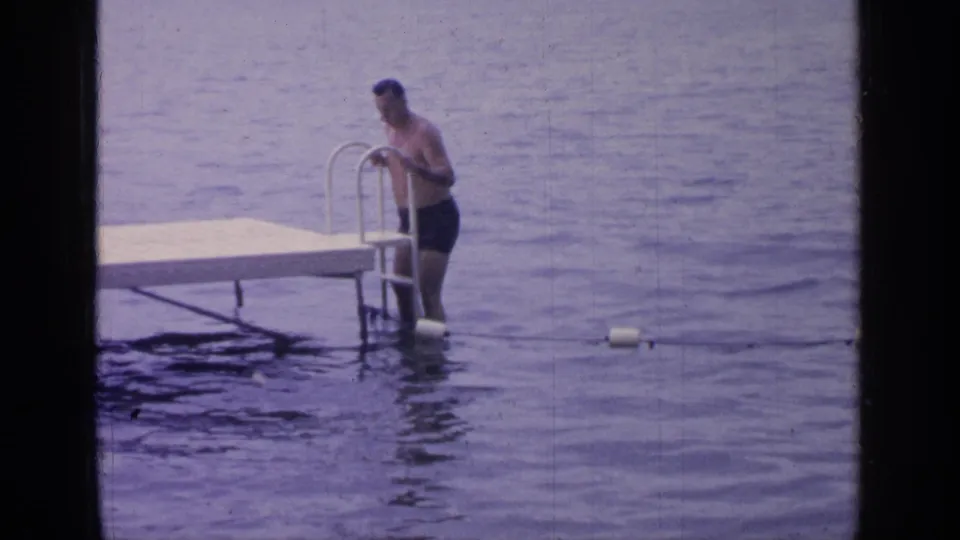At Greenwood Lake in 1964, the man grips the metal ladder beside the dock while standing knee-deep in rippling water.