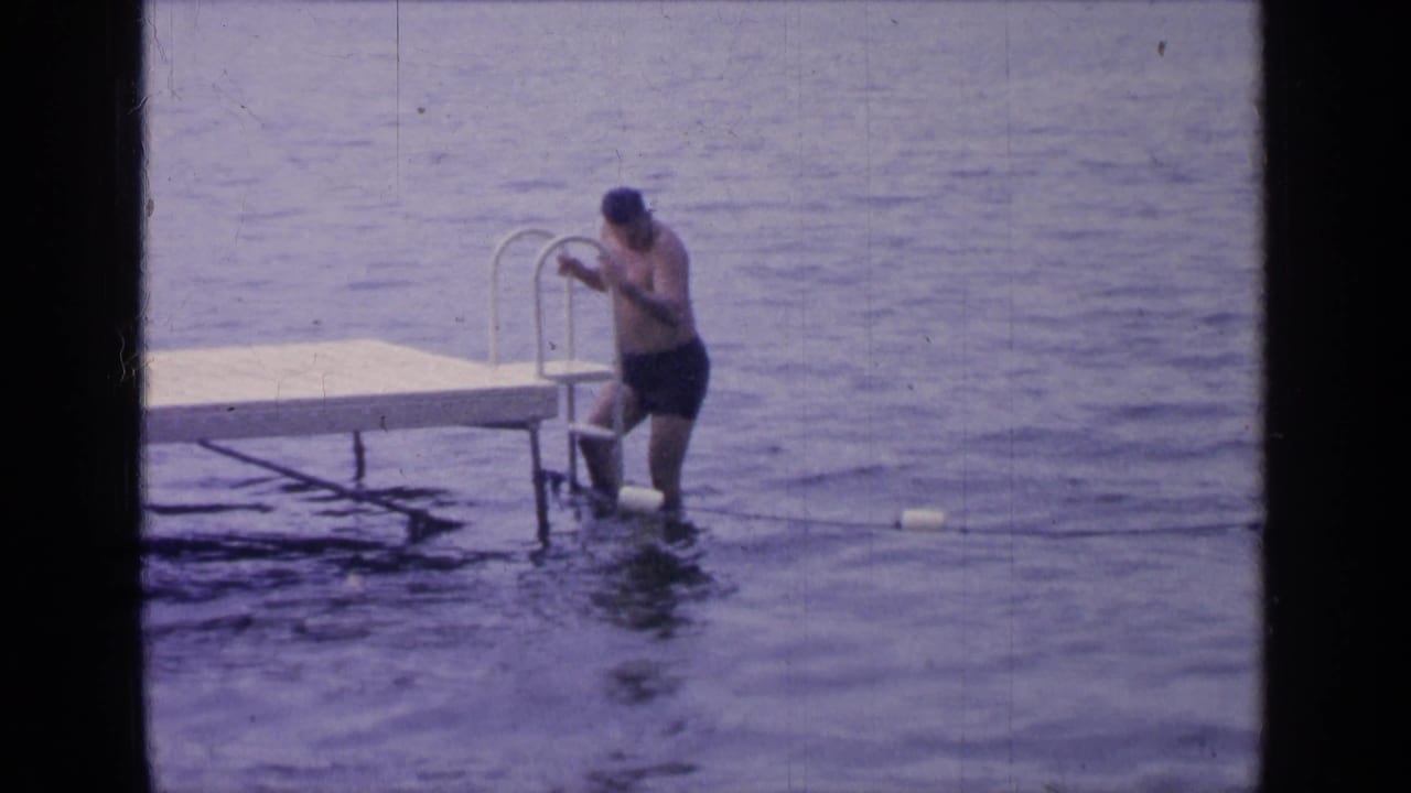 At Greenwood Lake in 1964, the man stands in the water beside the dock ladder as soft blue-purple 8mm tones cover the lake scene.