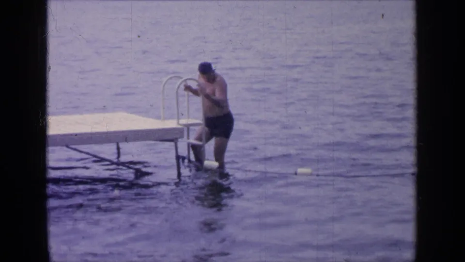 At Greenwood Lake in 1964, the man stands in the water beside the dock ladder as soft blue-purple 8mm tones cover the lake scene.