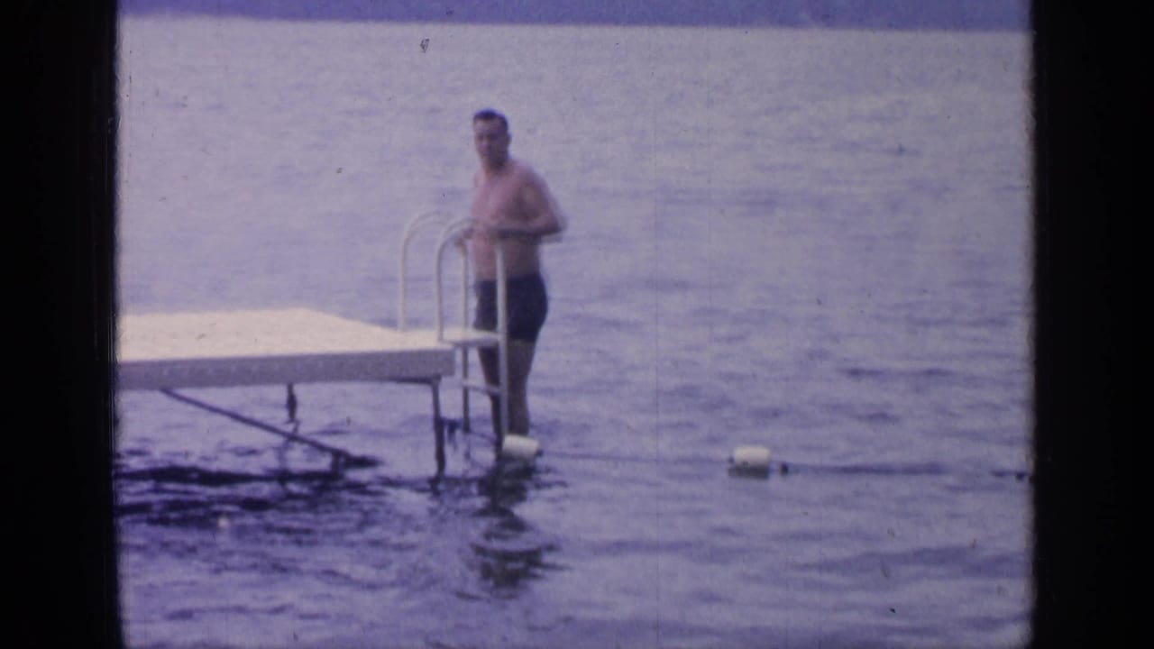 At Greenwood Lake in 1964, a shirtless man in dark swim trunks stands by a metal ladder at the edge of a wooden dock.