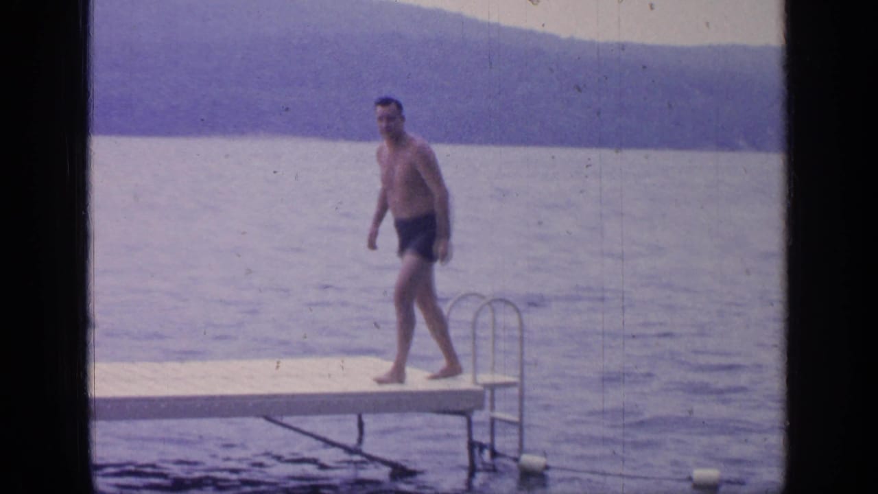 At Greenwood Lake in 1964, the man walks barefoot on the wooden dock with lake water and a wooded shoreline behind him.