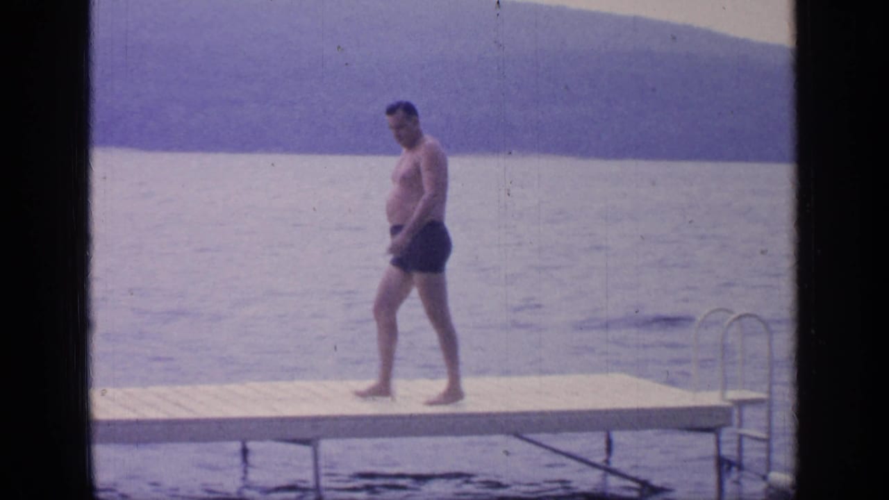 The 1964 Greenwood Lake home-movie frame shows the man walking along the dock with the ladder and lake surface nearby.