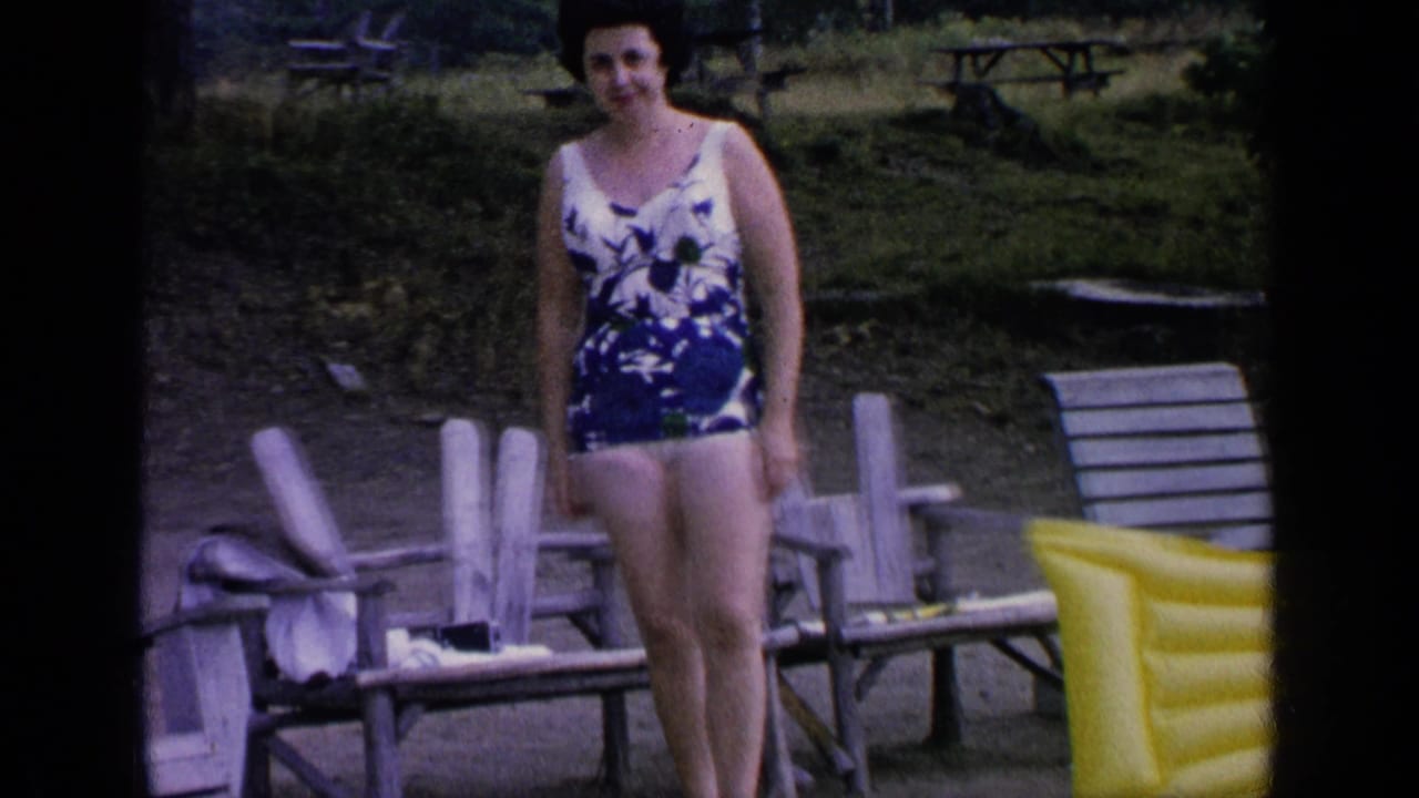 At Greenwood Lake in 1964, a woman in a blue and white floral one-piece swimsuit stands by wooden chairs near the water’s edge.