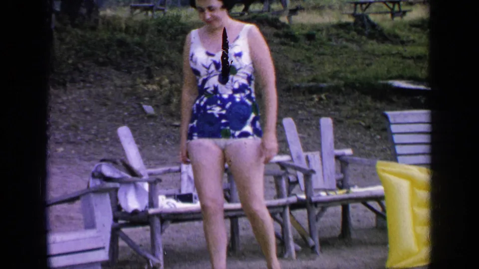 The woman in a blue floral swimsuit glances down as she stands near wooden chairs at Greenwood Lake, New York, in 1964.