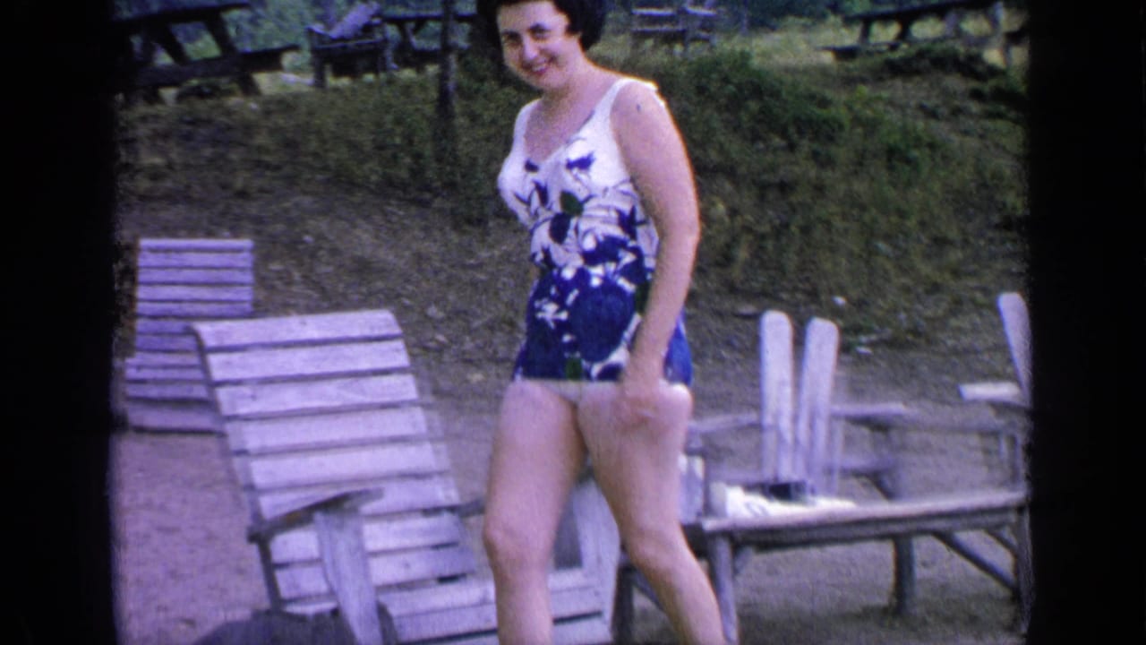 The woman smiles while walking past wooden lounge chairs in 1964 home-movie footage from Greenwood Lake, New York.