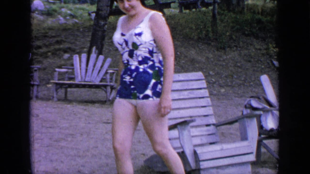 A woman in a blue floral swimsuit walks by wooden chairs at Greenwood Lake, with additional lakeside seating behind her.