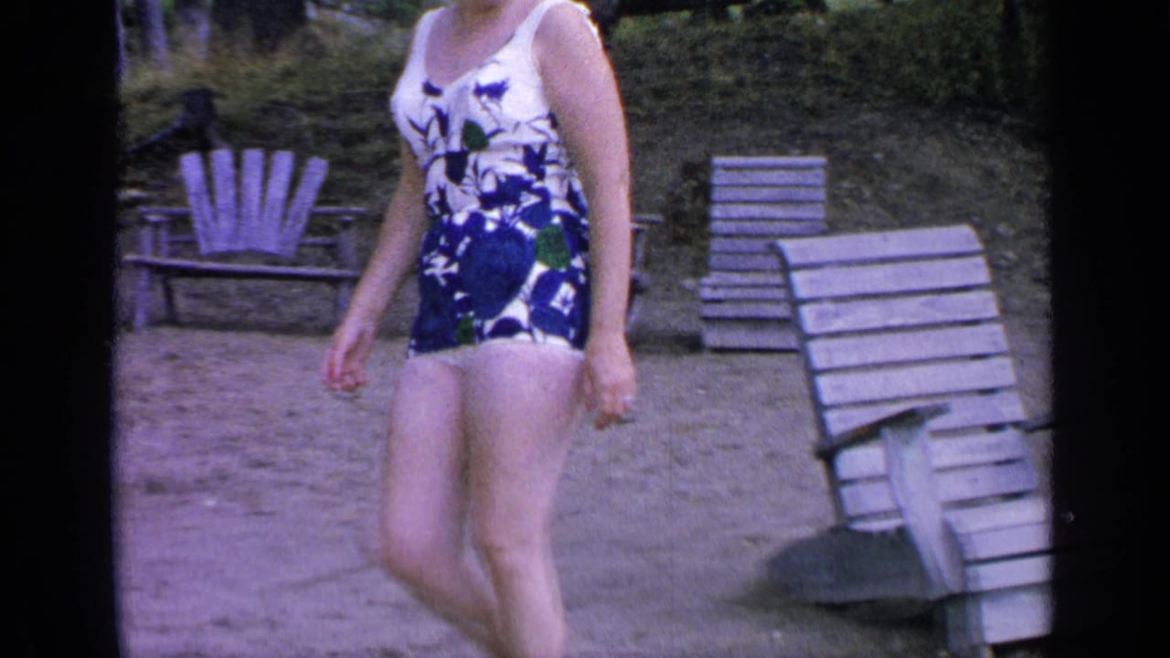 Woman in a floral one-piece swimsuit walking on sand near wooden beach chairs.