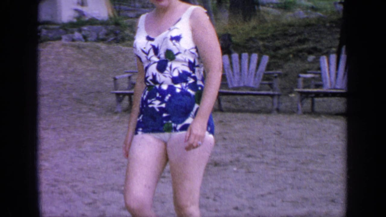 The woman stands near sandy lakeside seating at Greenwood Lake, wearing a blue and white floral one-piece swimsuit.