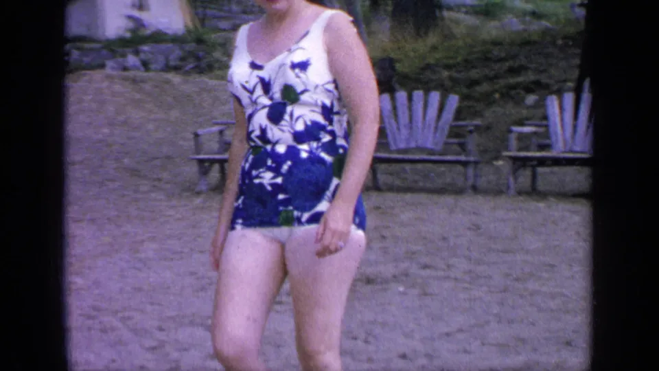 The woman stands near sandy lakeside seating at Greenwood Lake, wearing a blue and white floral one-piece swimsuit.