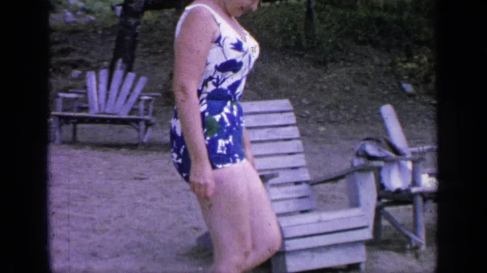 Side view of the woman walking across the sandy area near wooden lounge chairs in the 1964 Greenwood Lake home movie.