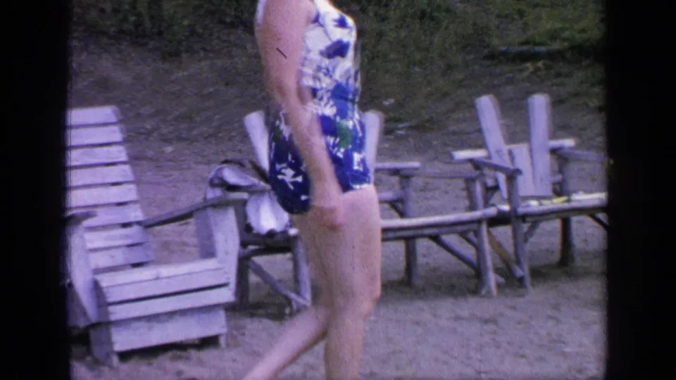 The woman continues past rows of wooden chairs on sandy ground, with trees and 8mm frame borders visible.